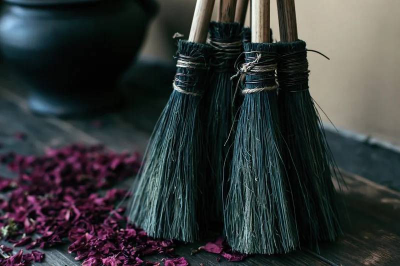 Four dark bristle brooms with wooden handles standing upright on a wooden surface beside dried rose petals and a black cauldron