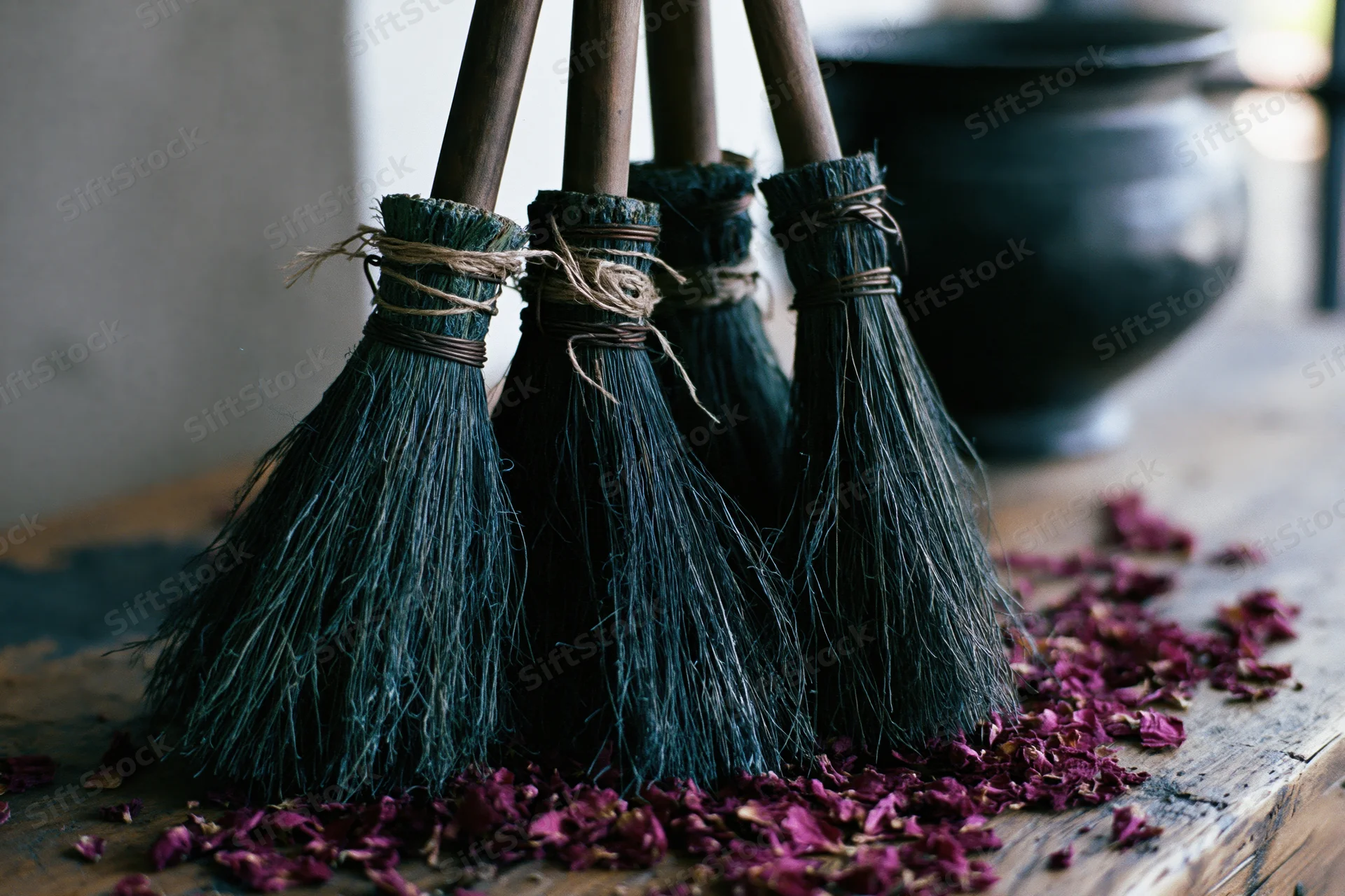Four dark bristle brooms with wooden handles standing upright on a wooden table scattered with rose petals near a black cauldron