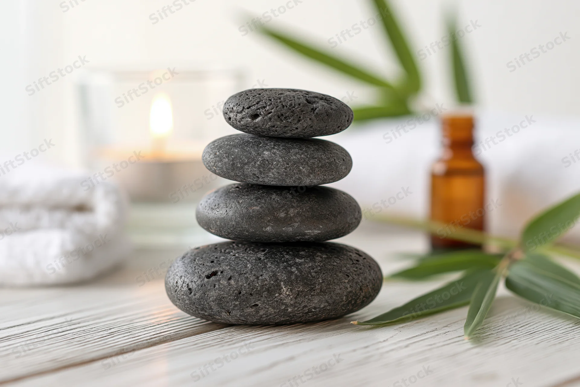 Stacked basalt stones on a wooden table with bamboo, candle, and essential oil bottle in a spa setting