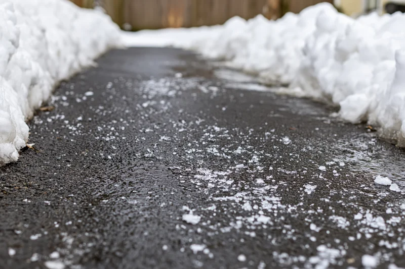 Cleared asphalt driveway with snowbanks on either side and scattered ice and slush on the surface