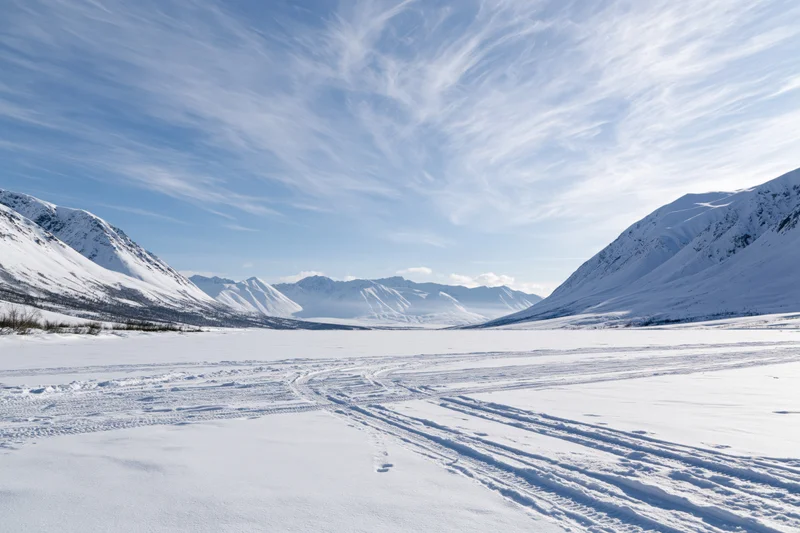 Snow-covered valley with tire tracks crossing frozen lake between snow-capped mountains under blue sky