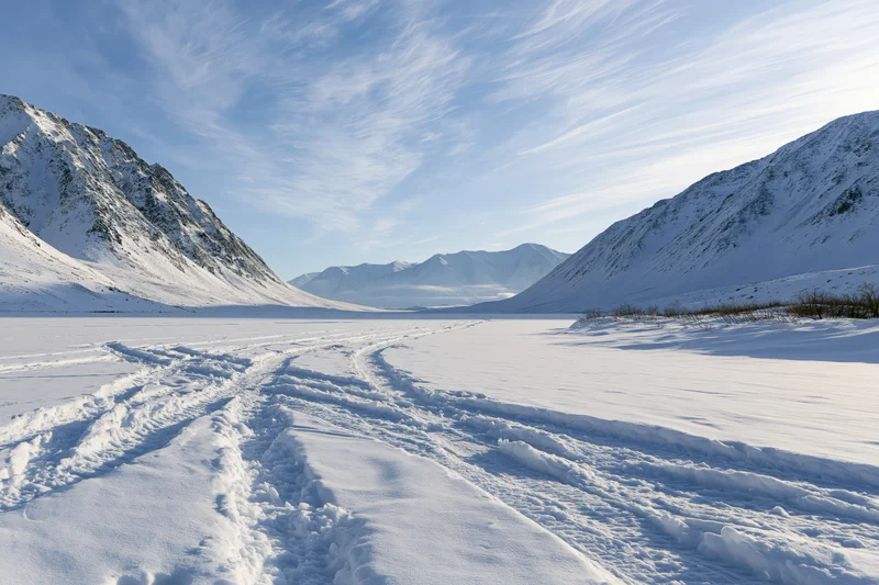 Tire tracks crossing a frozen lake between snow-covered mountains under a blue sky with wispy clouds