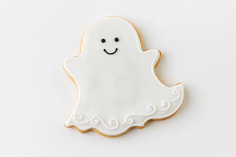 Ghost-shaped sugar cookie decorated with white icing and smiling face on white background