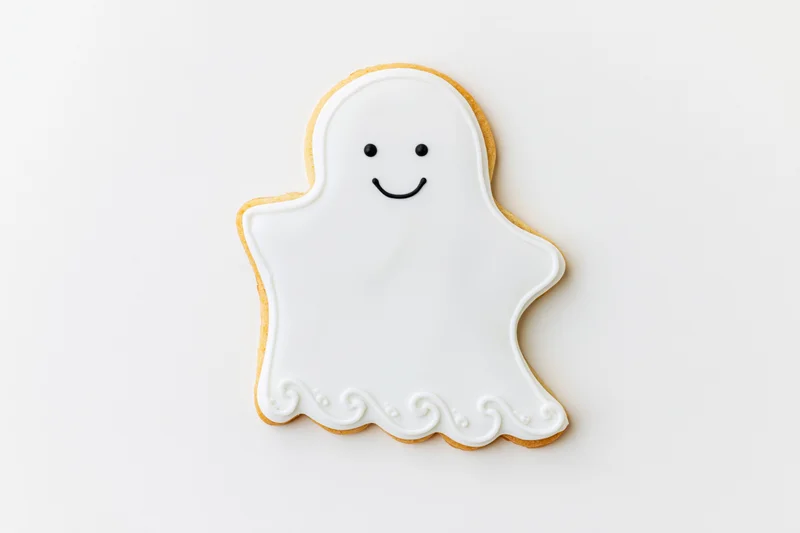 Ghost-shaped sugar cookie decorated with white icing and smiling face on white background