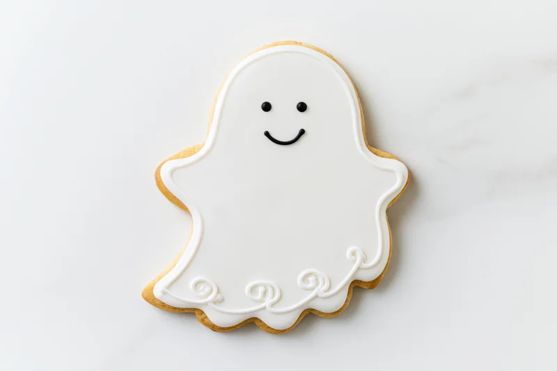 Ghost-shaped sugar cookie decorated with white icing and smiling face on marble surface