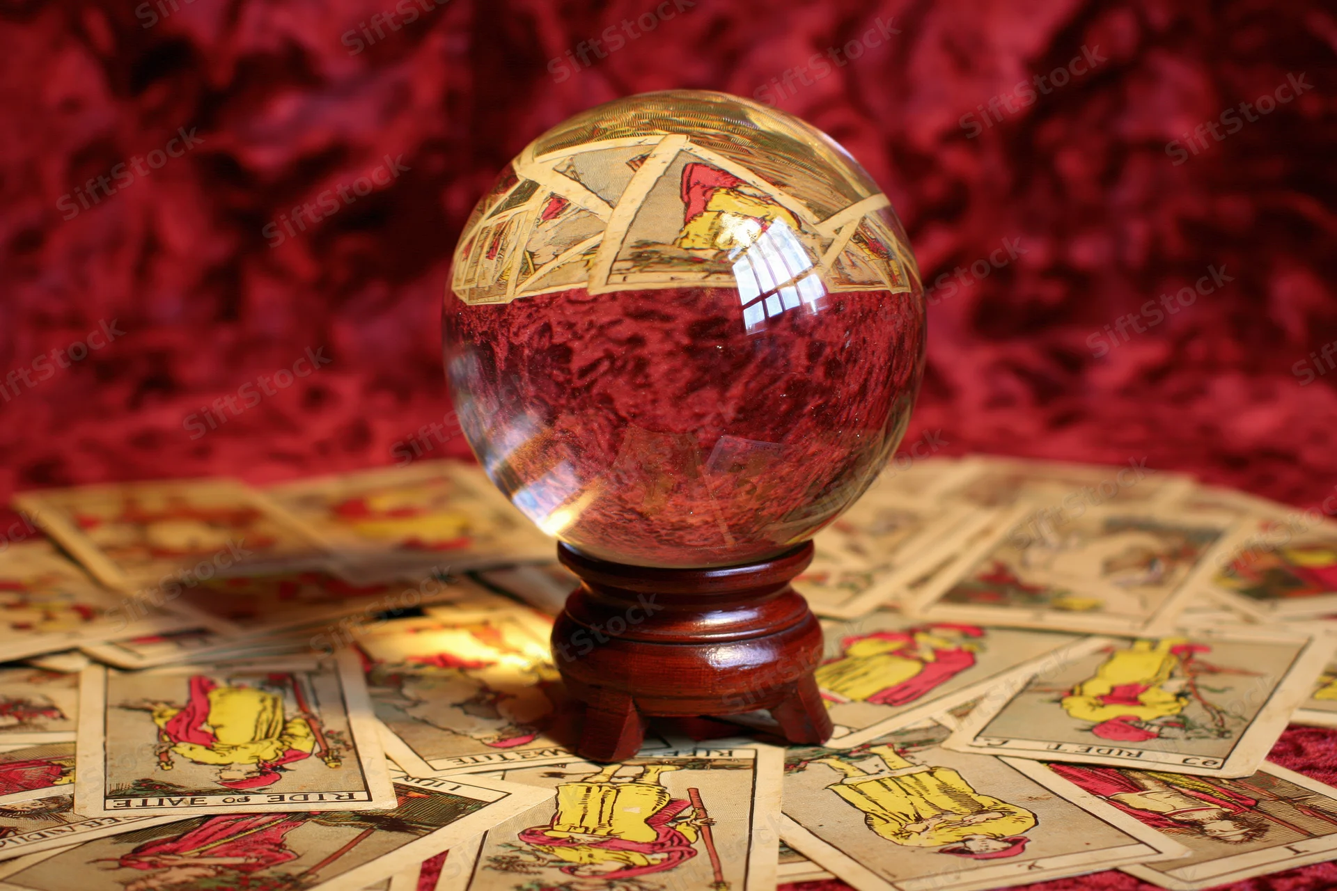 Crystal ball on wooden stand surrounded by scattered tarot cards on red velvet surface