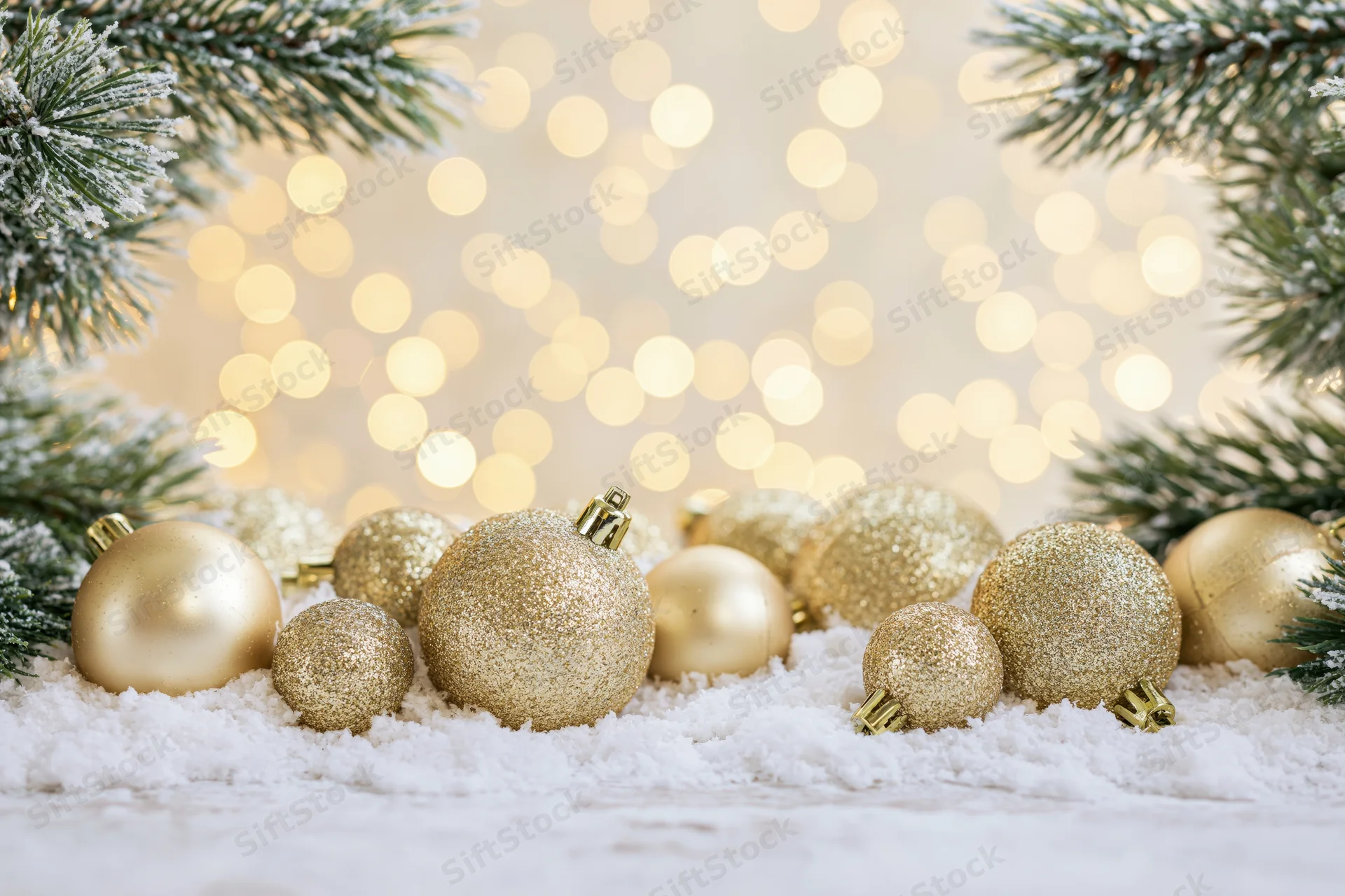 Gold christmas ornaments resting on snow with pine branches and bokeh lights in background
