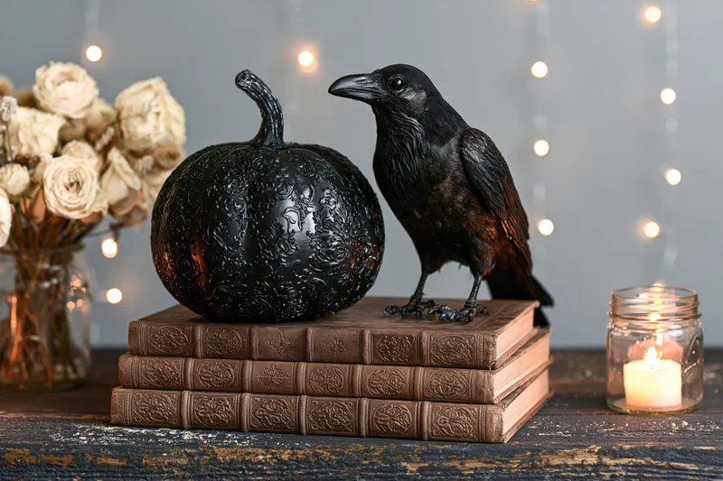 Black crow perched beside ornate black pumpkin on stack of vintage books with candle and dried roses on dark wood table
