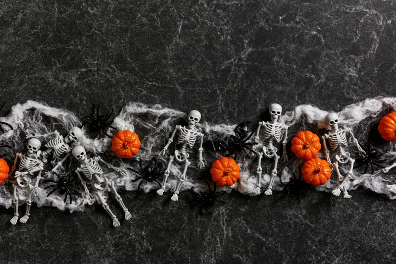 Halloween skeletons and pumpkins arranged along the bottom edge on a dark textured surface with cobwebs and spiders