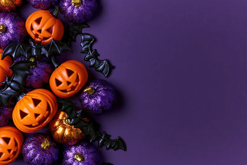 Halloween decorations including jack-o-lanterns and bats arranged in a flat lay on purple background with copyspace