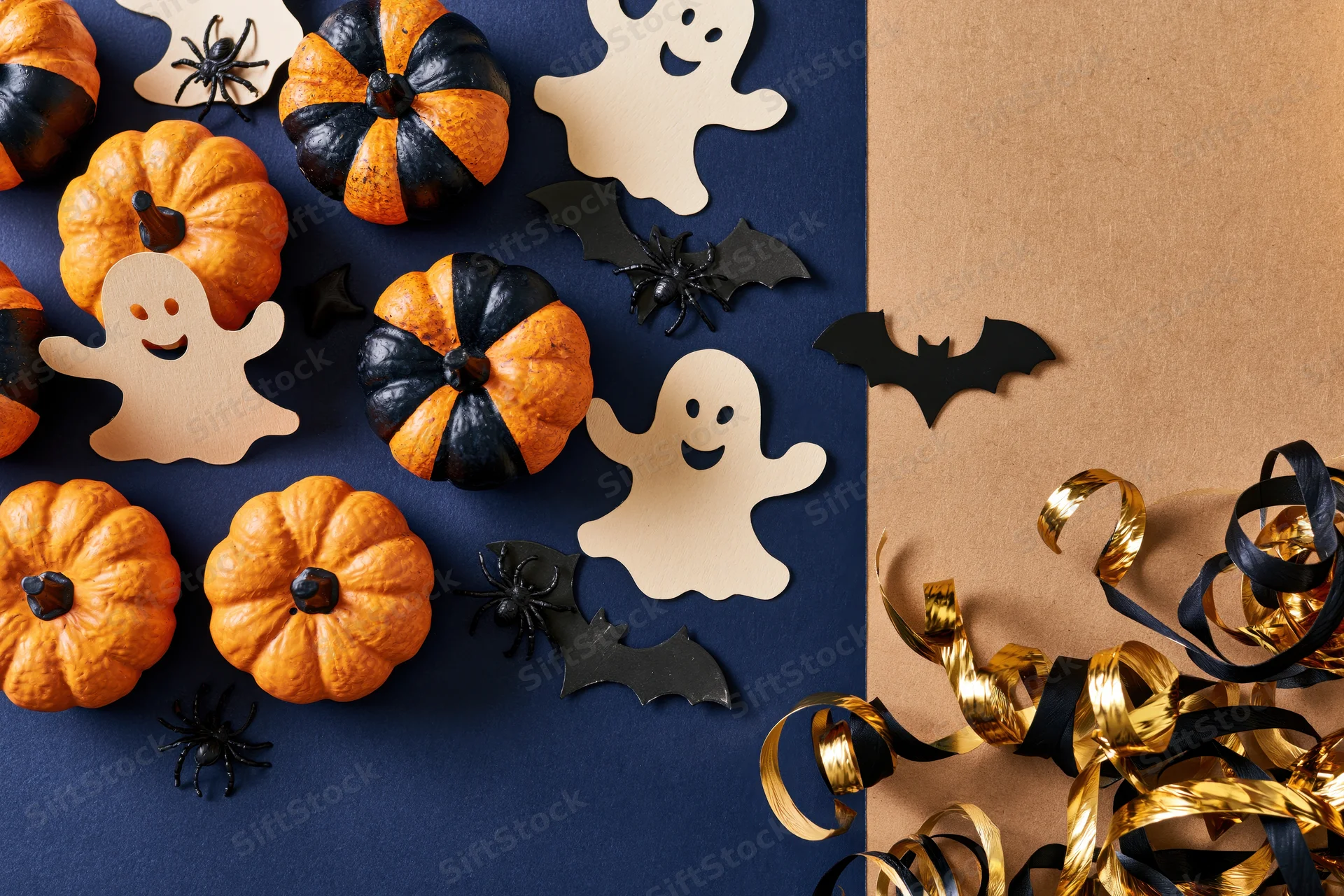 Halloween flat lay with orange and black pumpkins, paper ghosts, bats, spiders, and gold ribbons on dark blue and brown surface