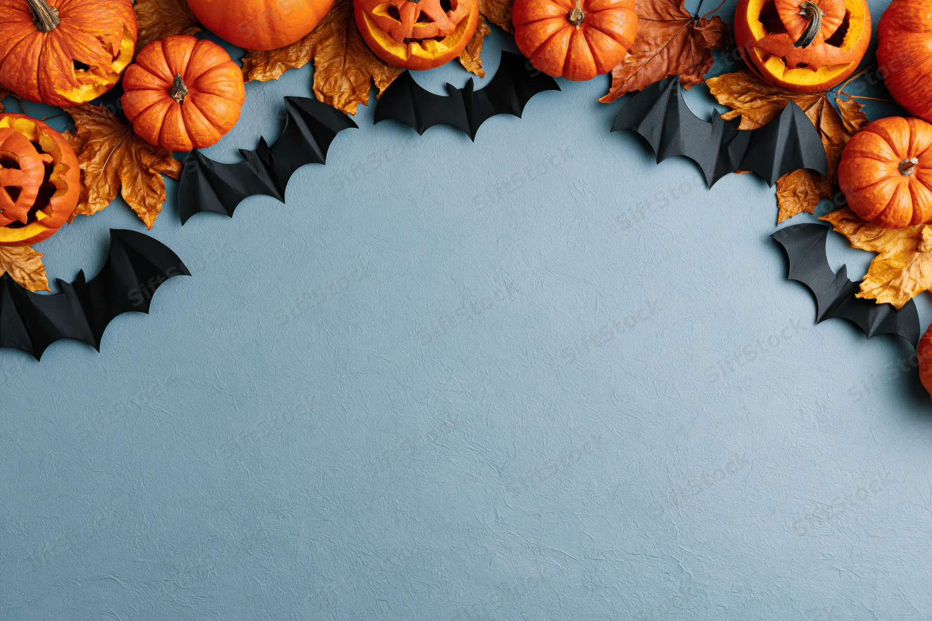 Halloween flat lay with carved pumpkins and paper bats arranged along the top edge on a blue textured surface