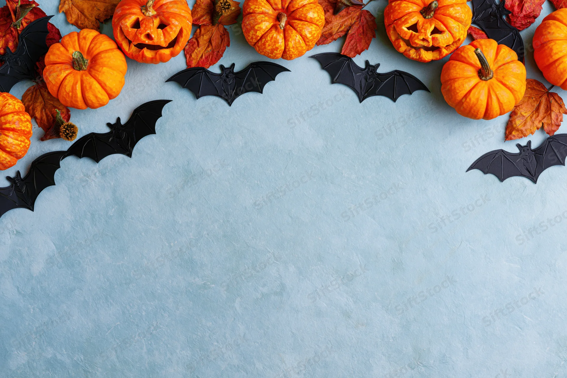 Halloween flat lay with carved pumpkins, bats, and autumn leaves arranged along the top edge of a blue surface, leaving copyspace below