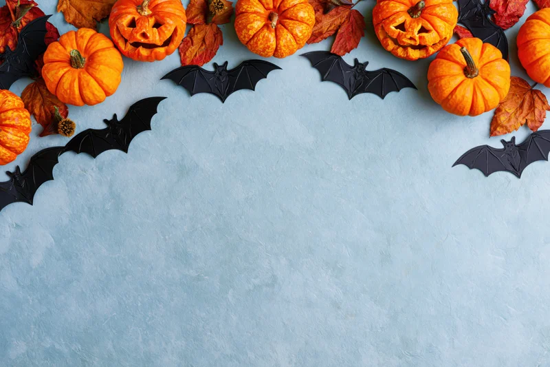 Halloween flat lay with carved pumpkins, bats, and autumn leaves arranged along the top edge of a blue surface, leaving copyspace below