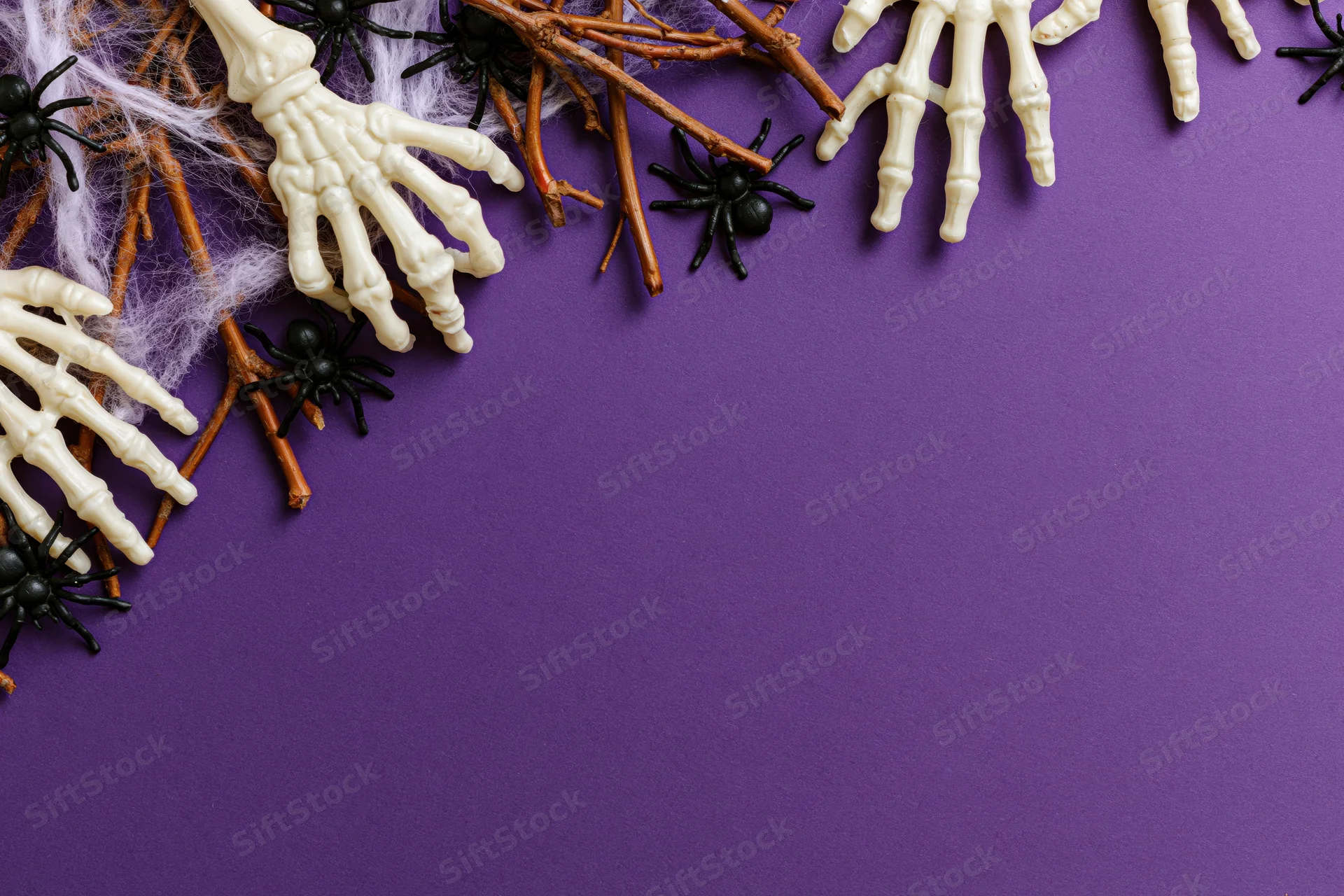 Plastic skeleton hands and spiders arranged with twigs and web on purple surface for halloween flat lay