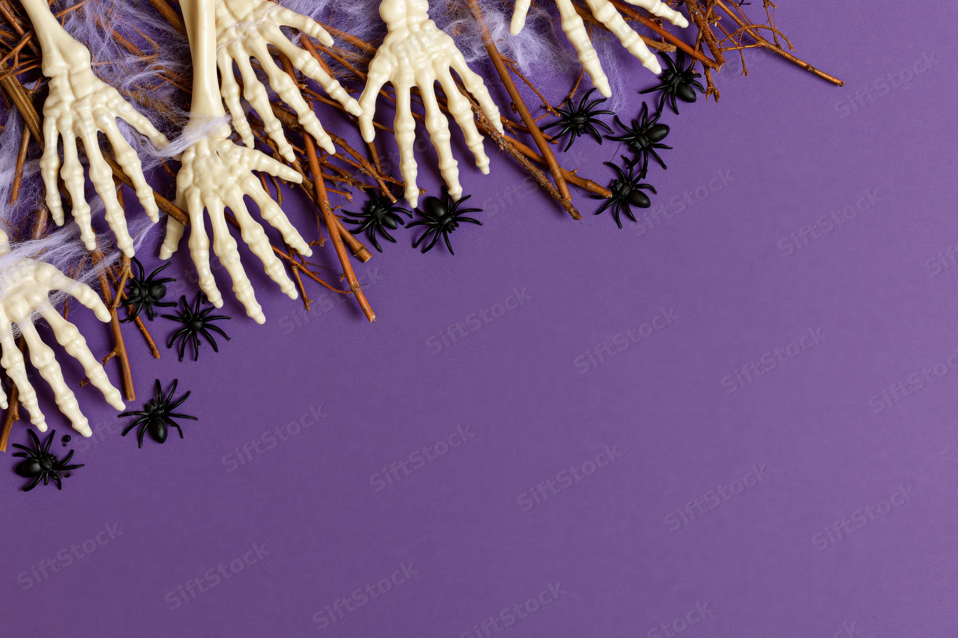 Plastic skeleton hands and black spiders arranged with twigs and web on purple background