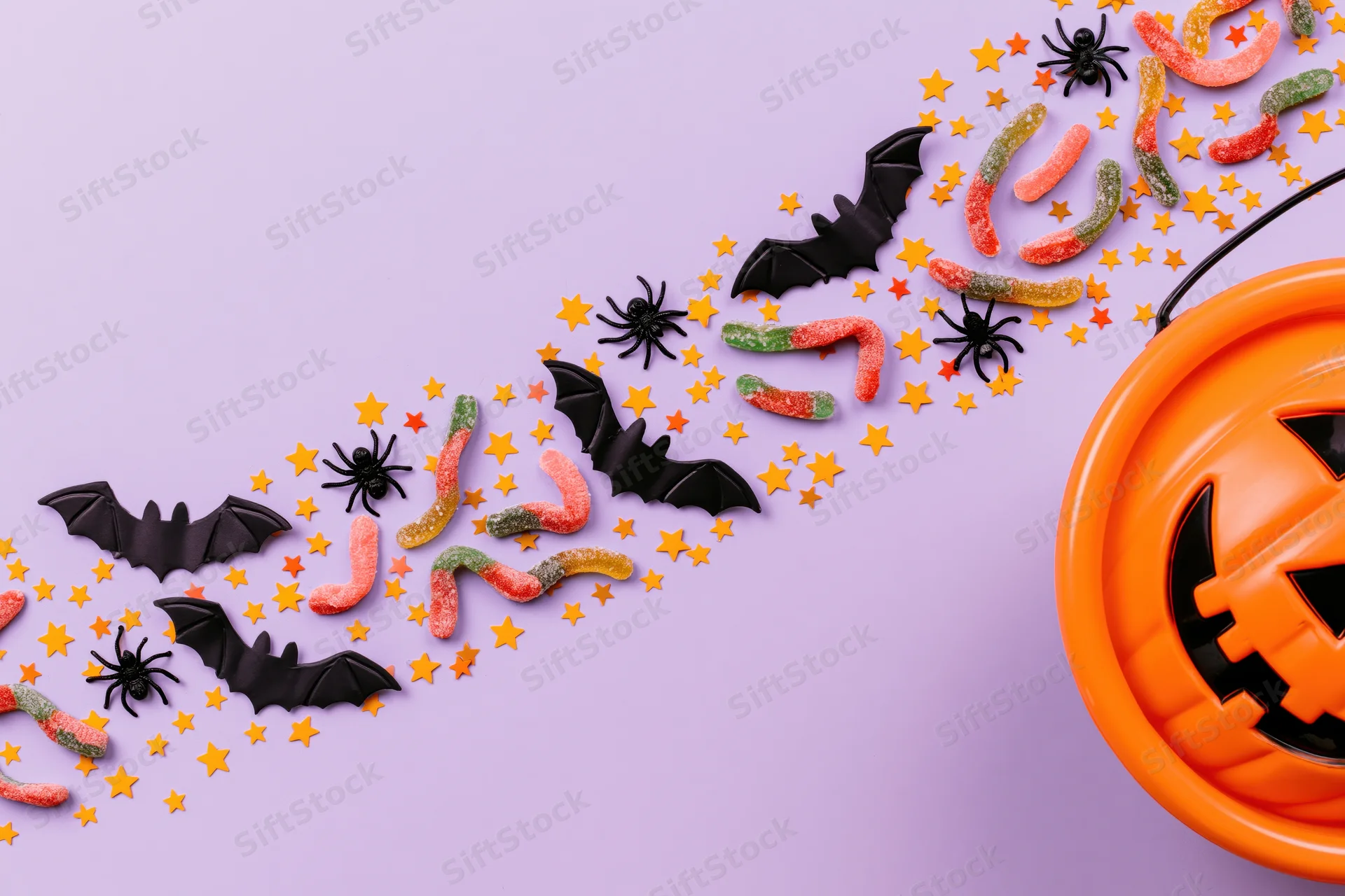 Orange plastic jack-o-lantern bucket with candy, bats, spiders and stars arranged in a diagonal flat lay on purple background