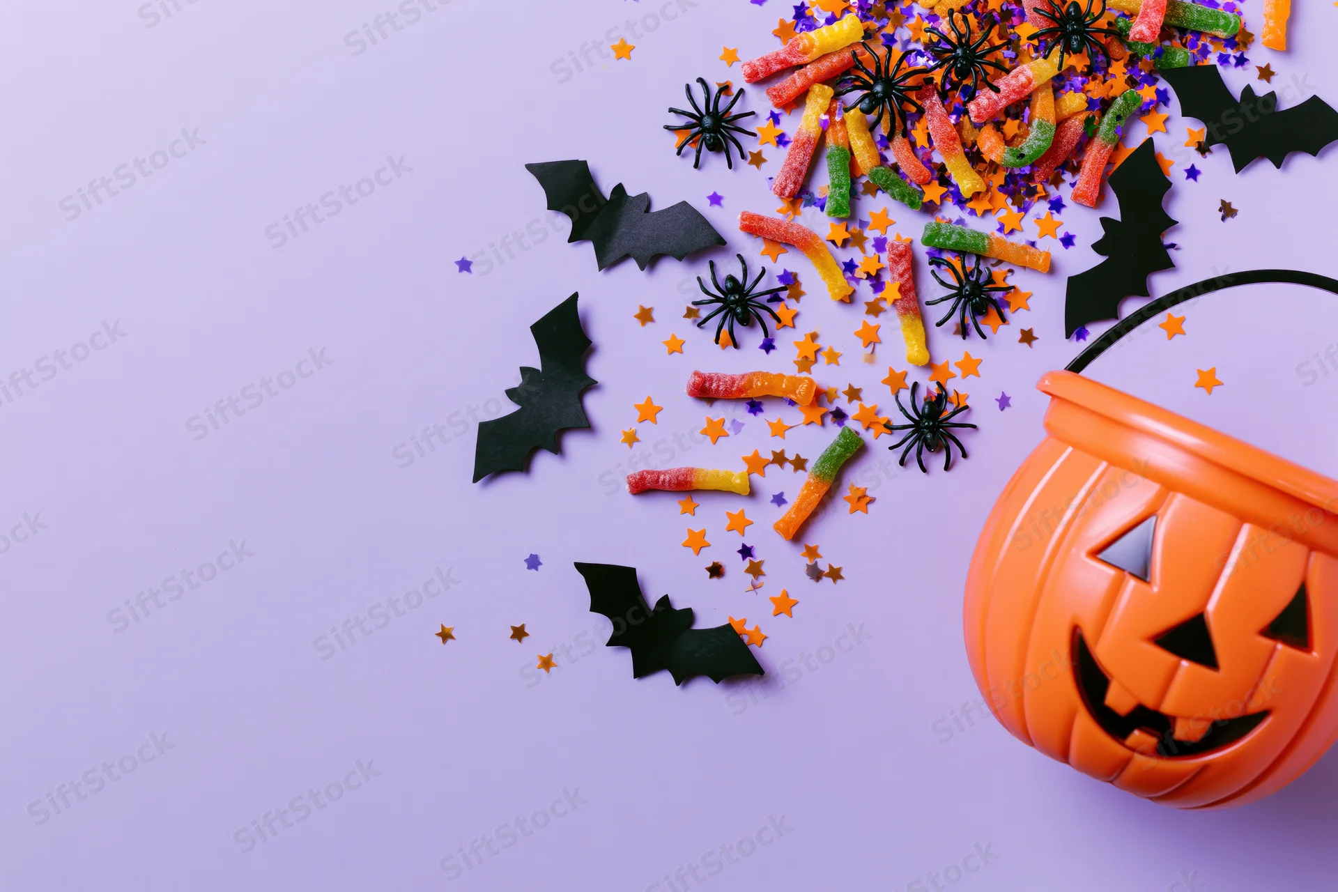 Orange plastic jack-o-lantern bucket with candy, spiders, bats and confetti on purple background
