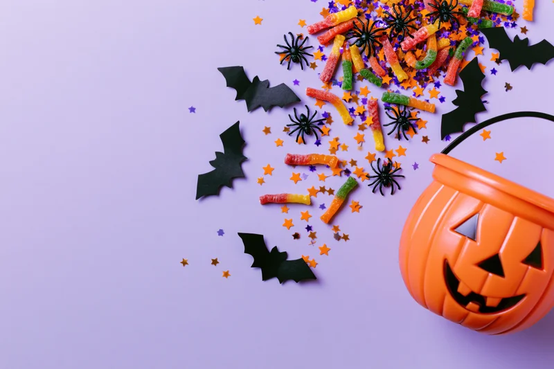 Orange plastic jack-o-lantern bucket with candy, spiders, bats and confetti on purple background