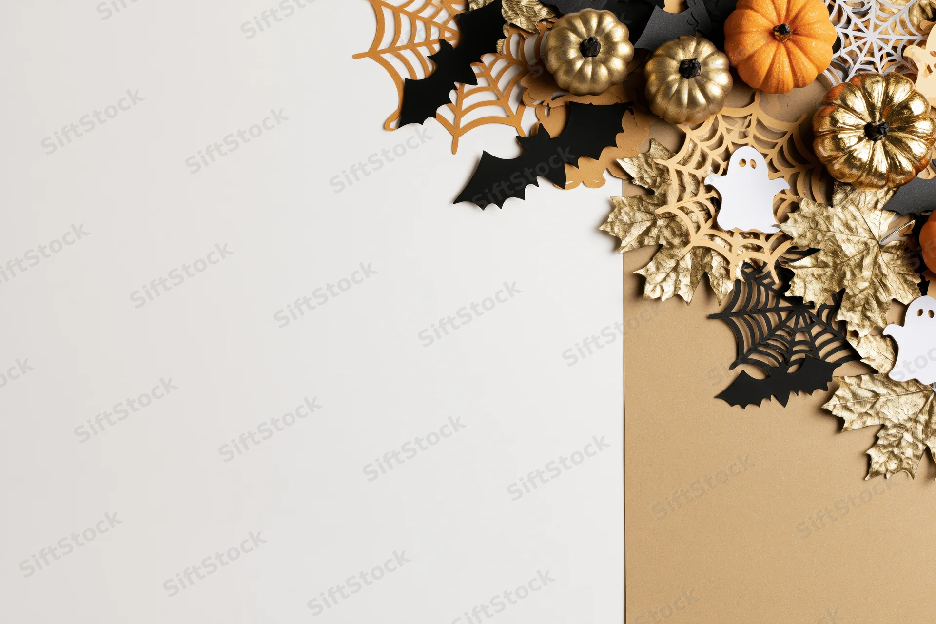 Halloween flat lay with paper bats, gold and orange pumpkins, spiderwebs, and leaves arranged on white and beige background
