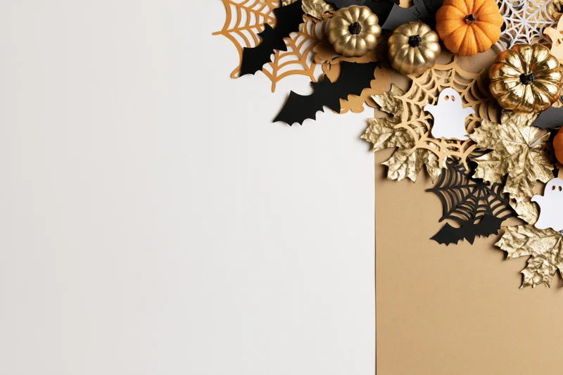 Halloween flat lay with paper bats, gold and orange pumpkins, spiderwebs, and leaves arranged on white and beige background