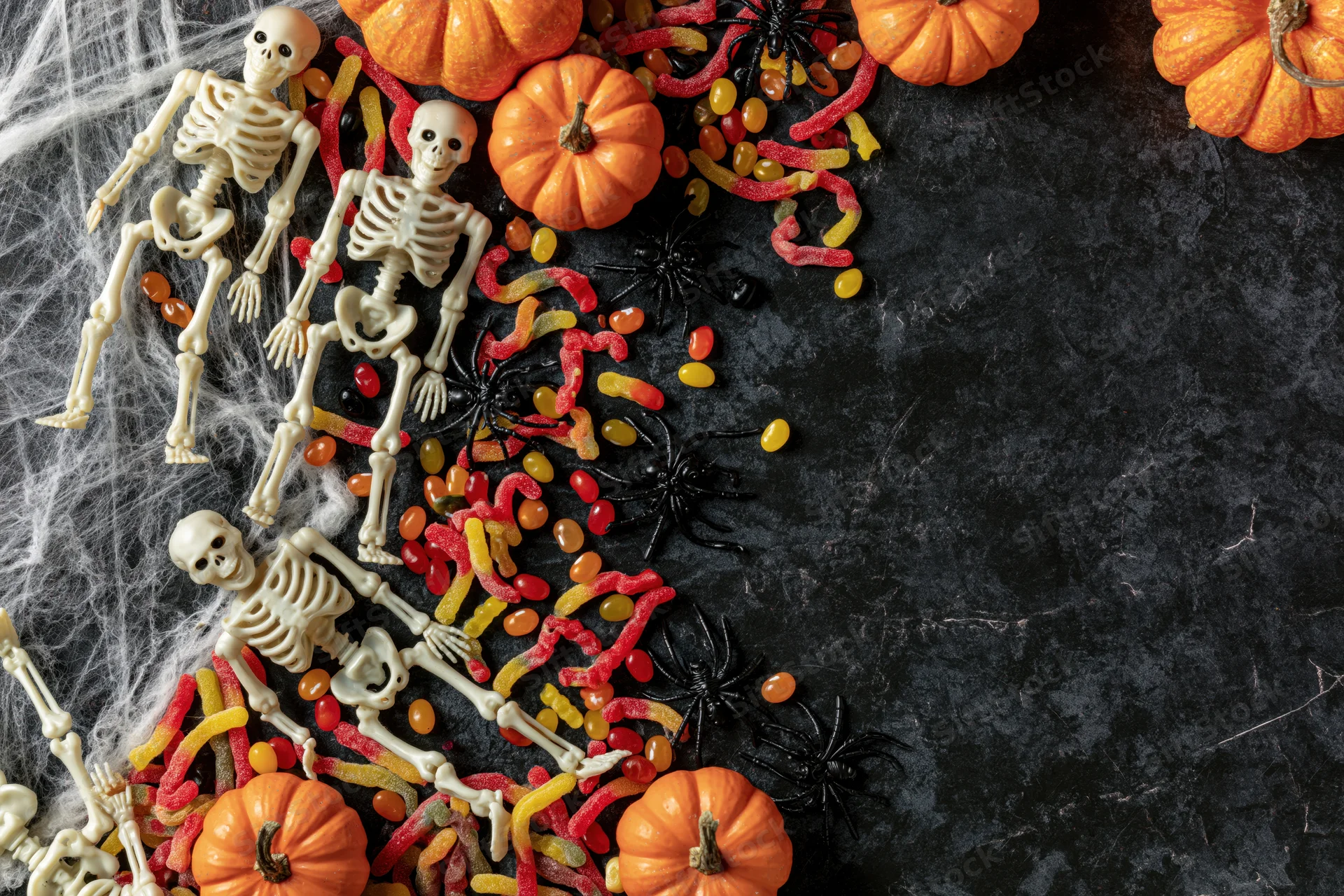 Plastic skeleton figurines and candy arranged with pumpkins and spiders on dark surface for halloween flat lay