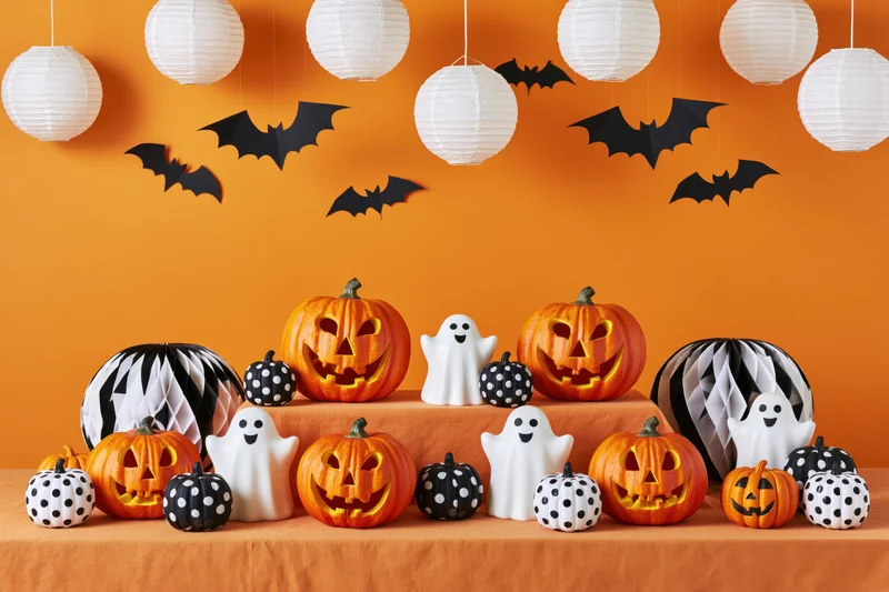 Halloween party table display with carved jackolanterns, ghosts, bats, and paper lanterns arranged on orange cloth against orange wall