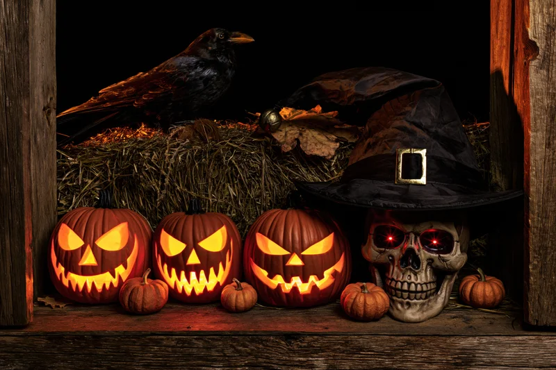 Carved pumpkins with glowing eyes and a skull in a witch hat arranged on a wooden barn doorstep with hay and a raven perched above