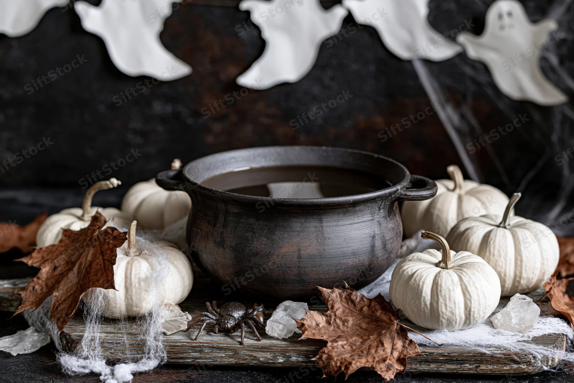 Black cauldron with white pumpkins and spider on weathered wooden board surrounded by cobwebs and autumn leaves for halloween decoration