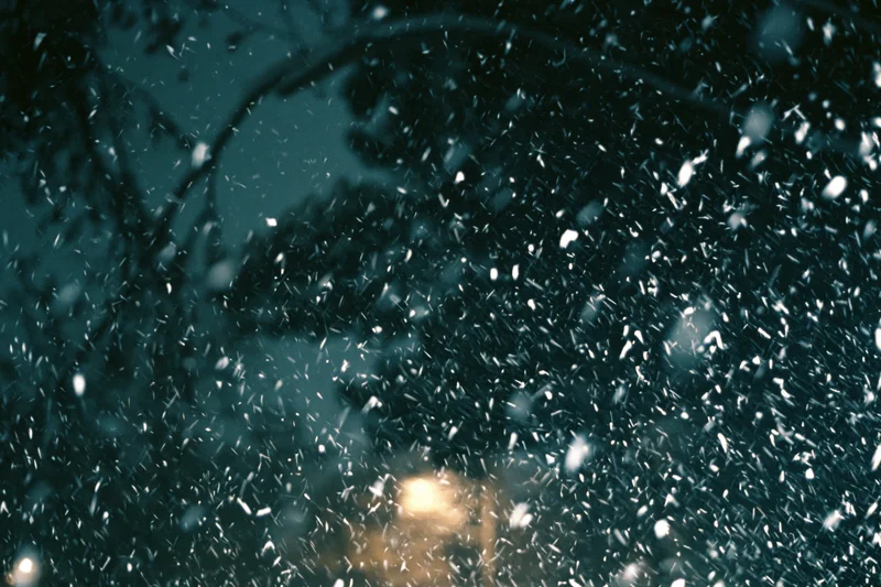Snow falling densely in a dark winter forest at night with blurred lights in the background