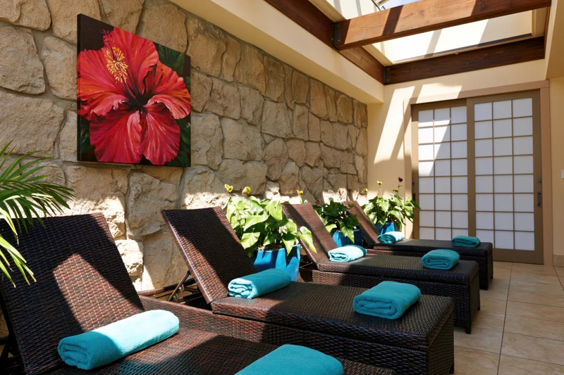Dark wicker lounge chairs with folded teal towels arranged in a stone-walled spa relaxation room with skylight and shoji door