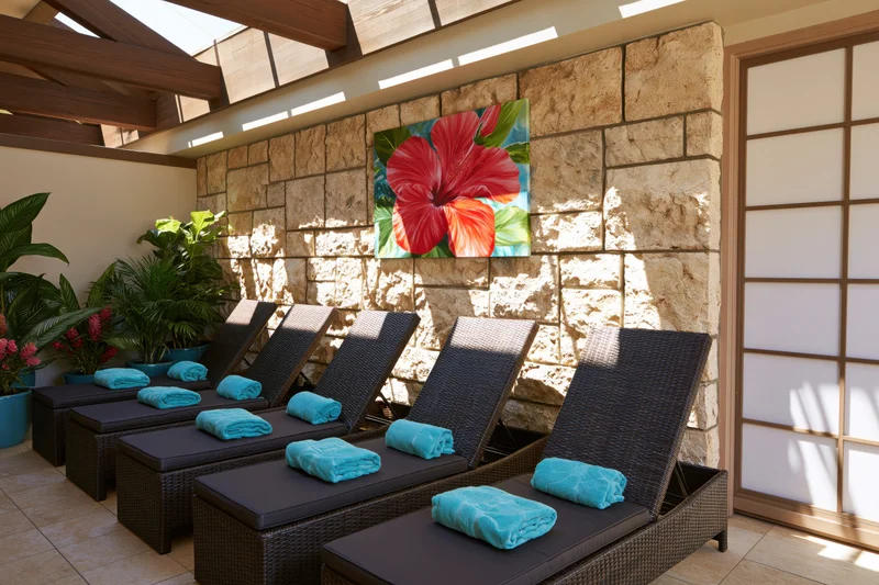 Dark wicker lounge chairs with turquoise towels arranged in a row under a stone wall with tropical artwork in a hotel spa relaxation room