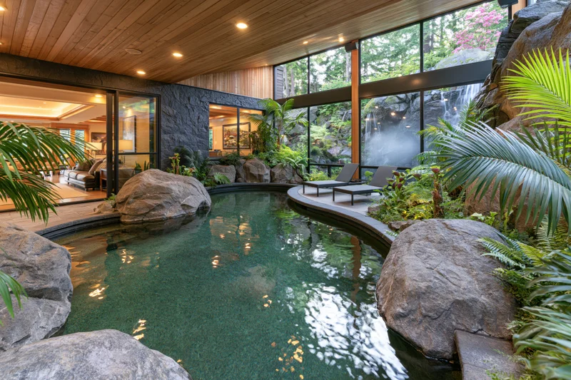 Curved indoor pool surrounded by rocks and tropical plants with glass walls overlooking a waterfall and lounge chairs nearby