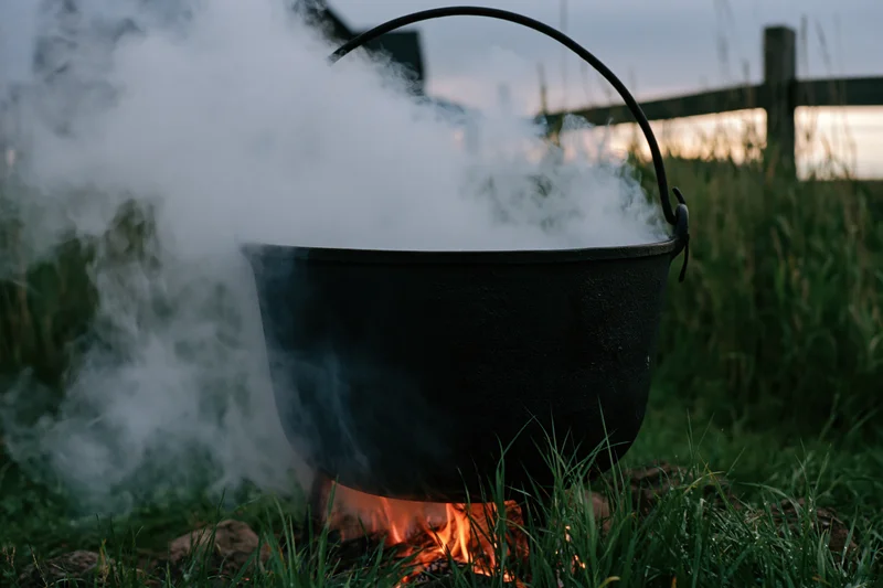 Black cast iron cauldron boiling over open fire in grassy field at dusk