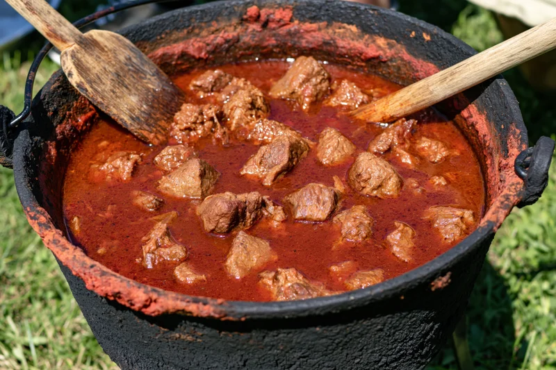 Deep red meat stew in a blackened cast iron cauldron outdoors with wooden spoons