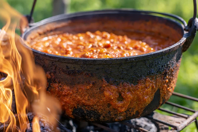Large blackened cast iron cauldron simmering orange-red stew over an open campfire outdoors