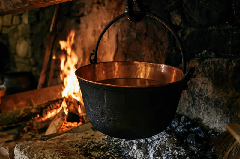 Blackened copper cauldron hanging over a stone fireplace with flames below