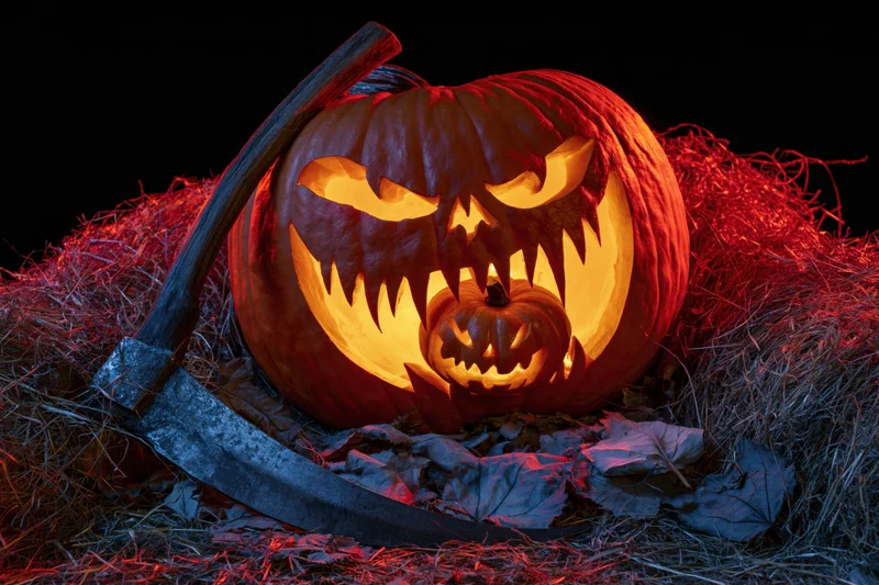 Large carved jack-o-lantern with menacing grin glowing in hay surrounded by axe and autumn leaves