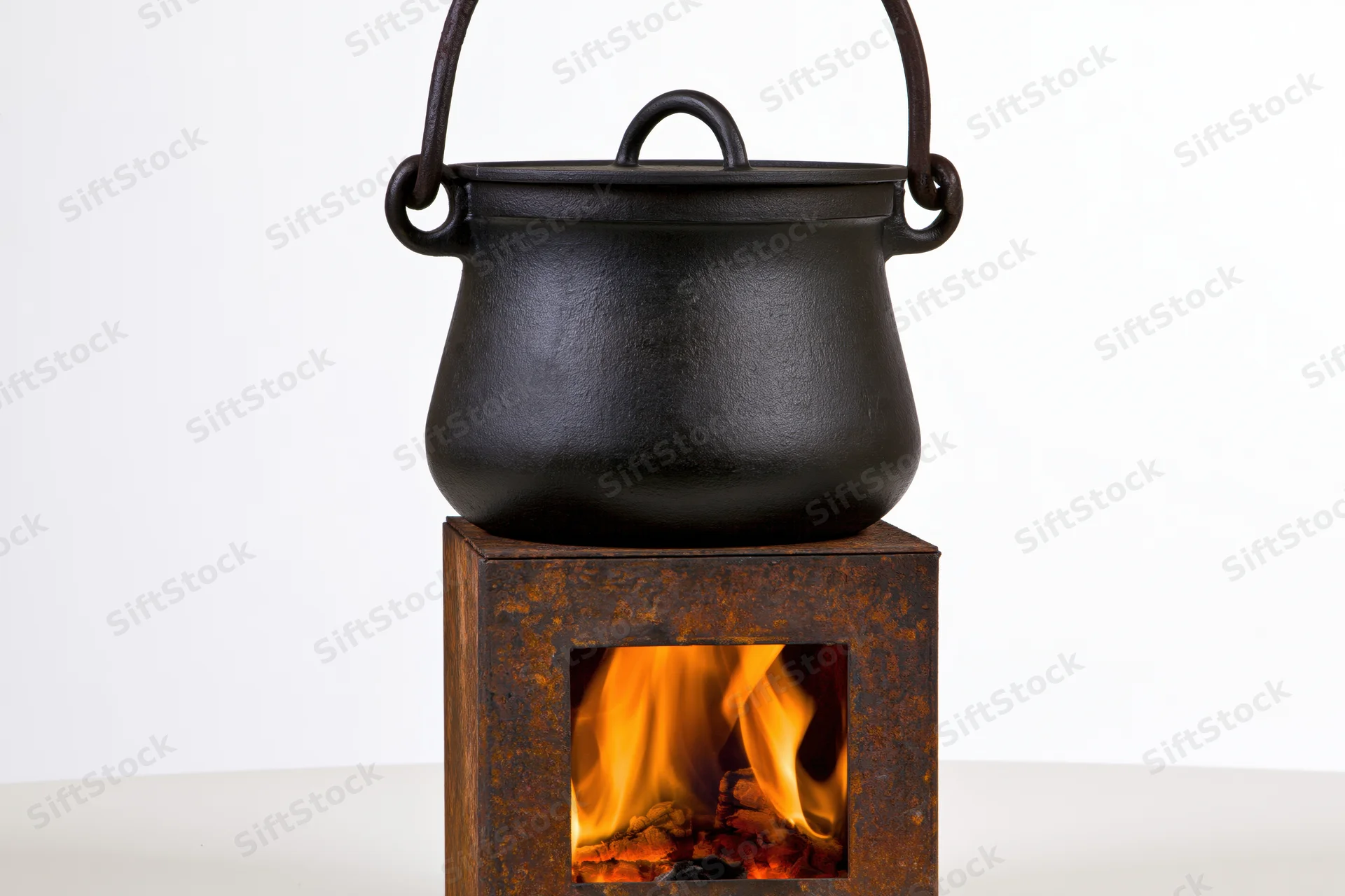 Black cast iron cauldron with curved bail handle resting on a rusted metal firebox with visible flames, isolated on white