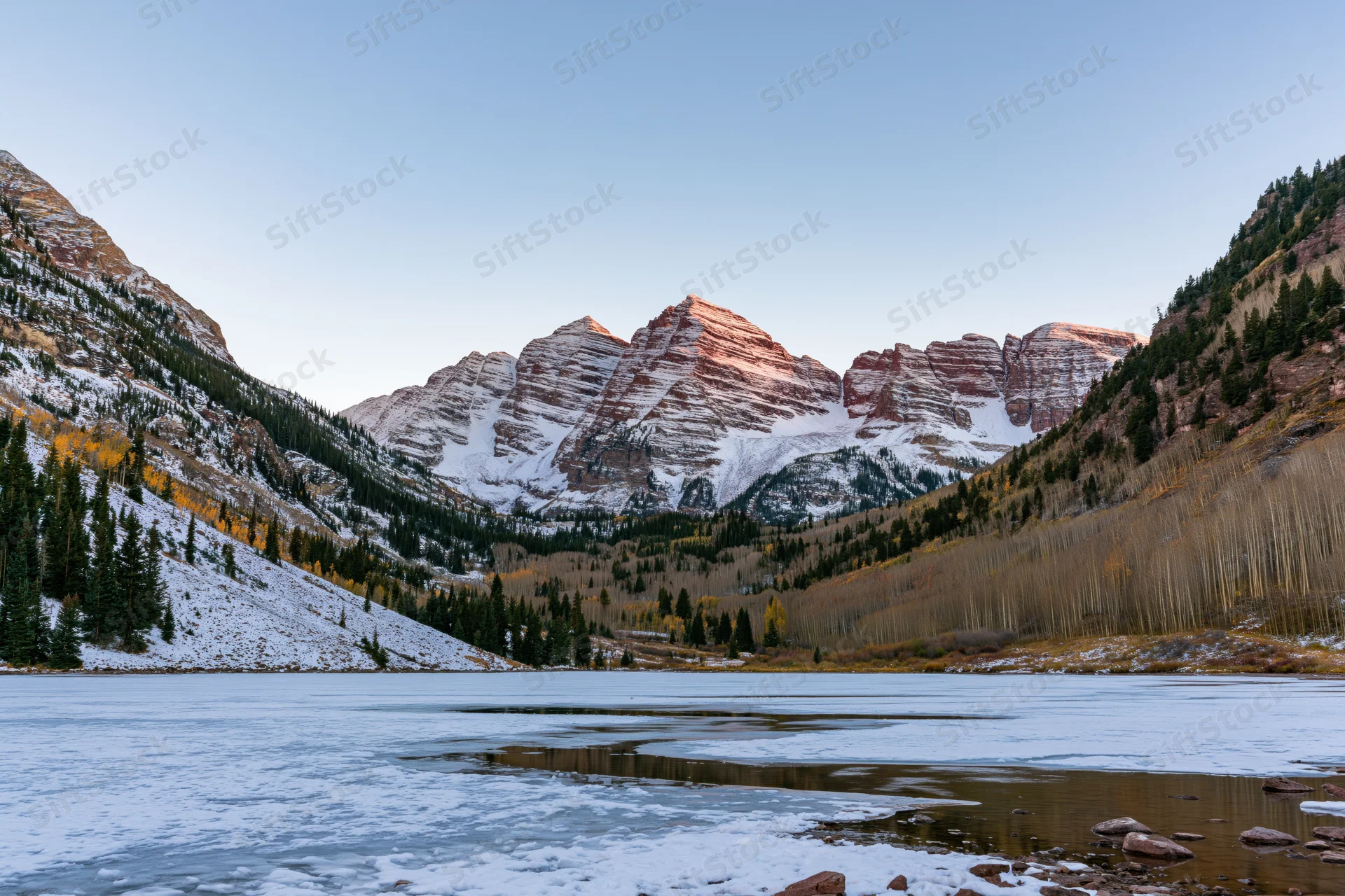 Snow-covered Maroon Bells peaks rising above a frozen lake in a mountain valley with autumn trees and evergreens