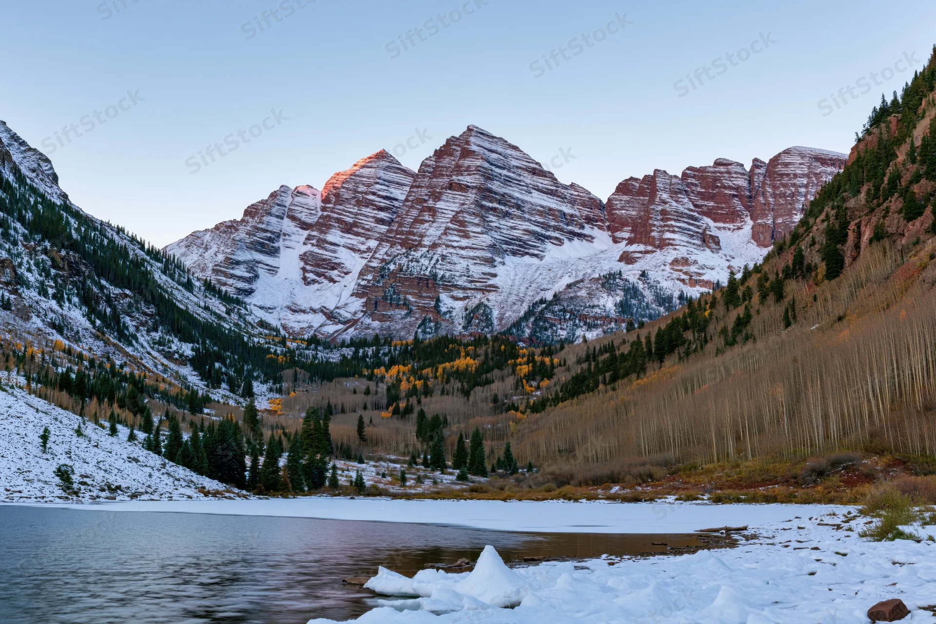 Snow-covered maroon bells peaks rising above a frozen lake in a mountain valley at dawn