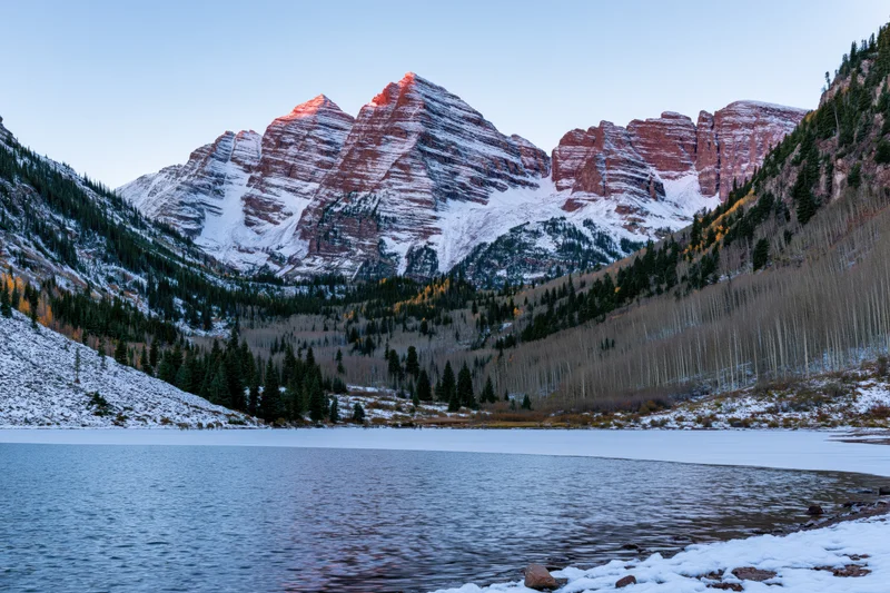 Snow-covered Maroon Bells peaks rising above a calm alpine lake surrounded by evergreen forest and aspen trees