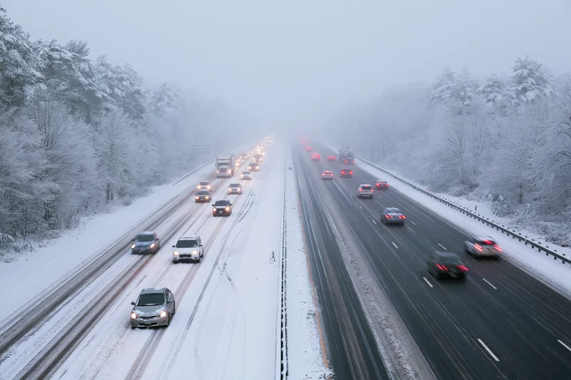 Multi-lane highway with slow-moving cars during heavy snowstorm, flanked by snow-covered trees