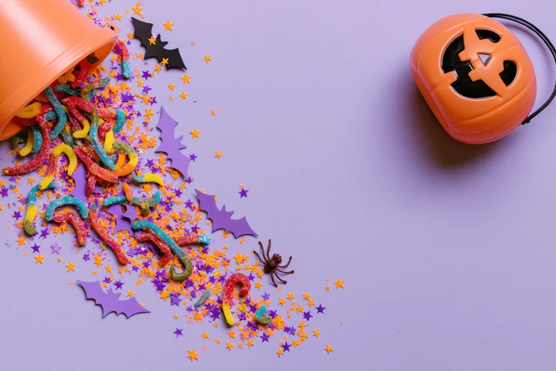 Orange plastic jack-o-lantern bucket tipped over with candy and decorations spilling onto purple surface