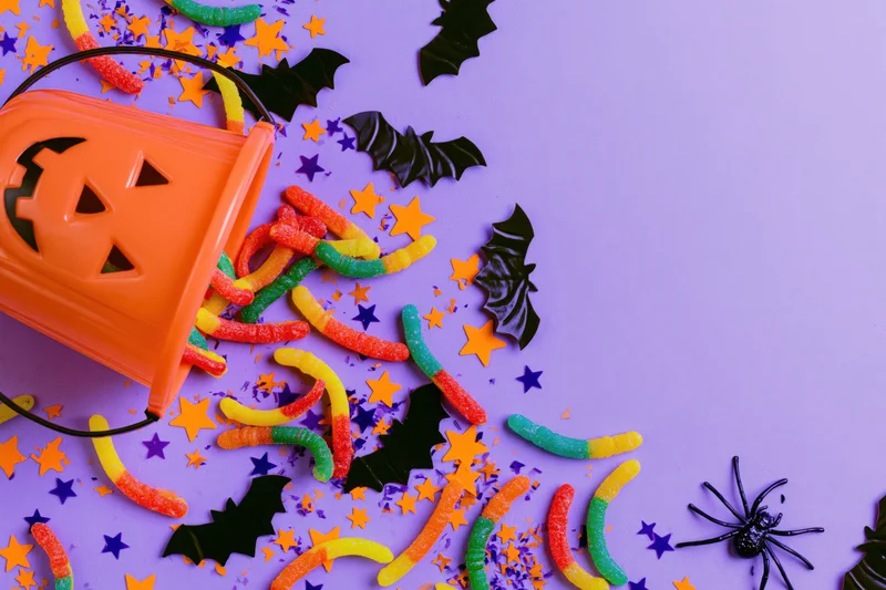 Orange plastic jack-o-lantern bucket tipped over with gummy worms and halloween decorations on purple background