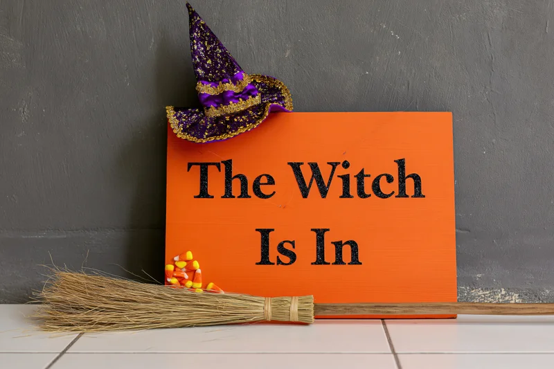 Orange halloween sign reading the witch is in with witch hat and broom on tiled floor against gray wall
