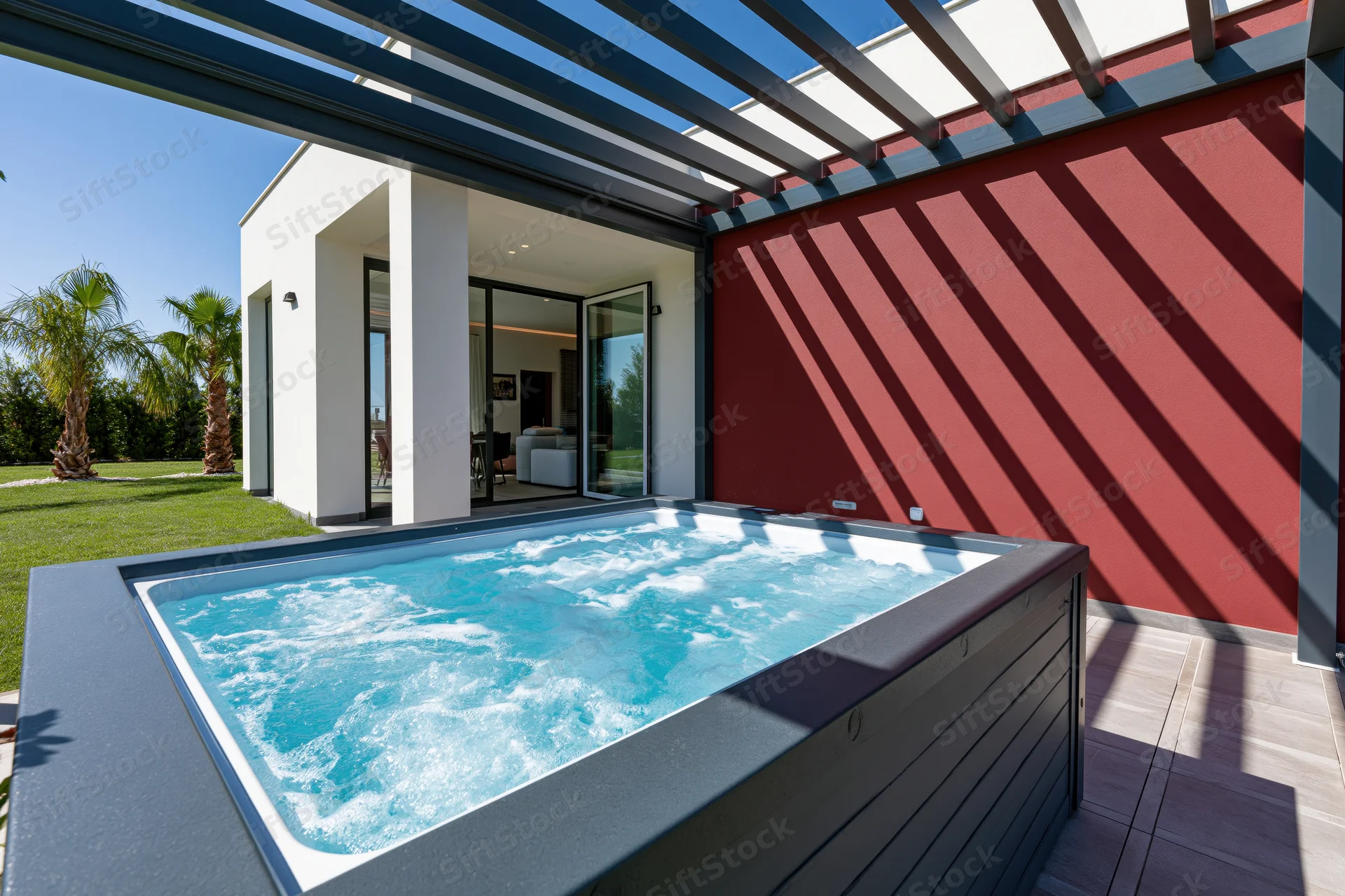 Bubbling hot tub on tiled patio under pergola with red wall and glass door view to interior