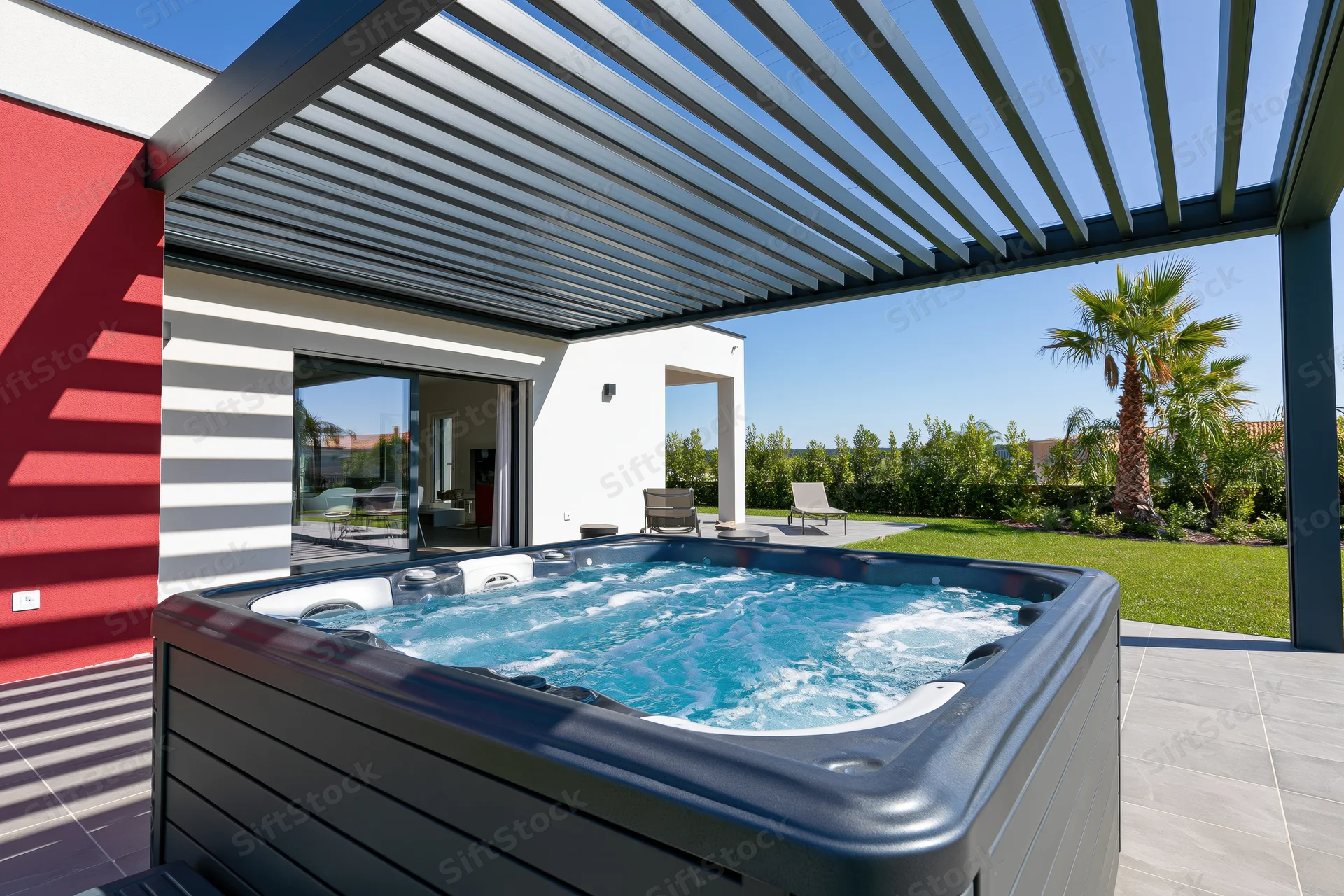 Bubbling hot tub on tiled patio under pergola beside modern house with garden view
