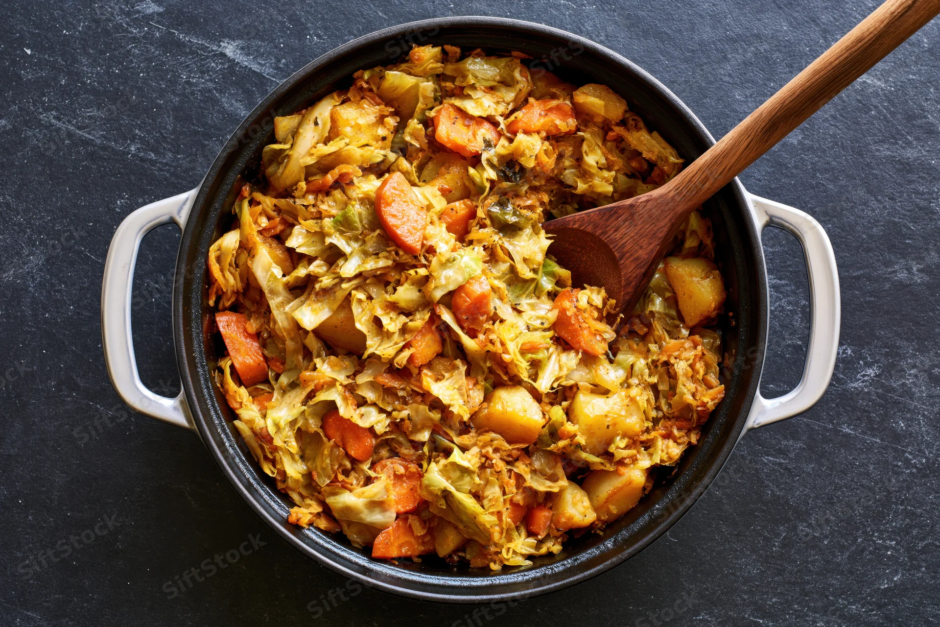 Sauteed cabbage and vegetables in a black cast iron pot with wooden spoon, overhead view on dark surface