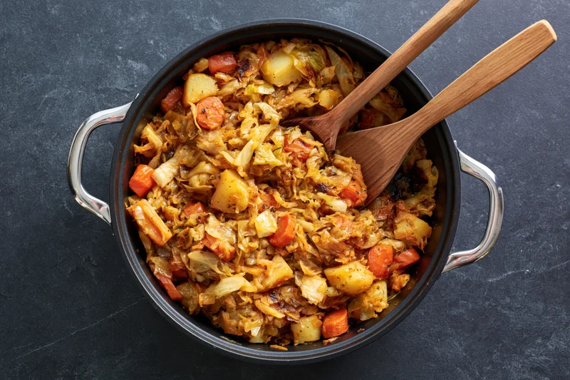 Sauteed cabbage and vegetables in a black cast iron pot with wooden spoons on dark surface